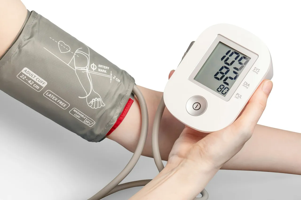 A person's arm wrapped in a grey blood pressure cuff while a hand holds a digital monitor showing a reading