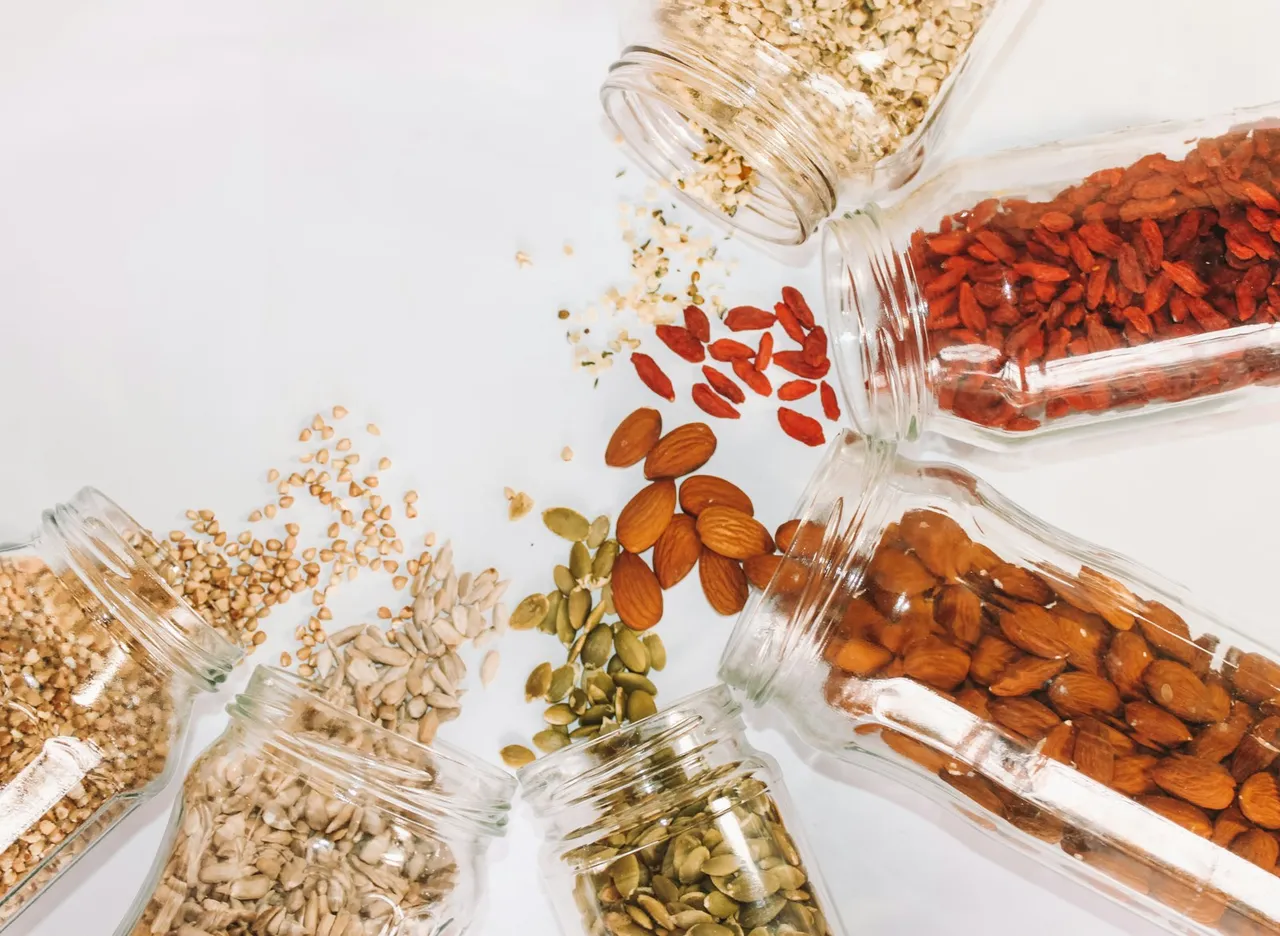 Glass jars of almonds, pumpkin seeds, goji berries and whole grains spilling onto a white surface