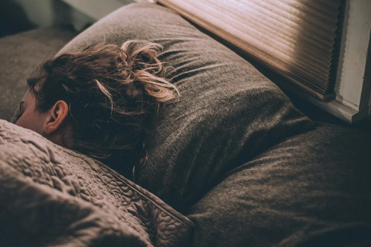 A person sleeping face-down on a pillow in a dim bedroom with morning light coming through blinds